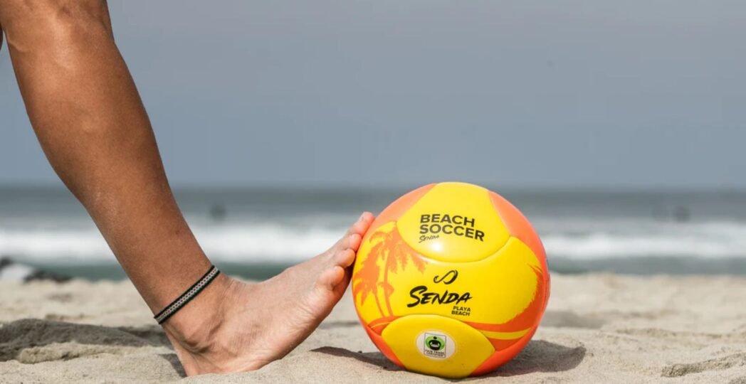 Person playing beach soccer with yellow and orange ball on sandy shore