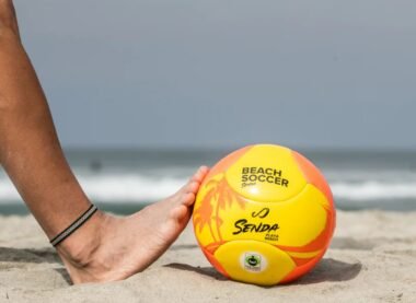 Person playing beach soccer with yellow and orange ball on sandy shore