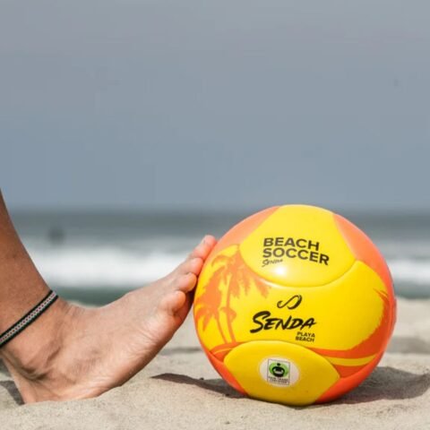Person playing beach soccer with yellow and orange ball on sandy shore